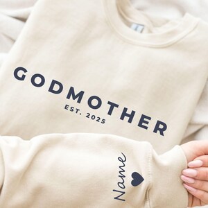 Pu&ograve; includere: Una felpa beige con la scritta blu navy "GODMOTHER" e "EST. 2025". La manica mostra un nome e un simbolo a forma di cuore. La felpa &egrave; piegata.