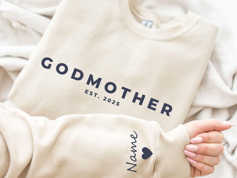 Pu&ograve; includere: Una felpa beige con la scritta blu navy "GODMOTHER" e "EST. 2025". La manica mostra un nome e un simbolo a forma di cuore. La felpa &egrave; piegata.