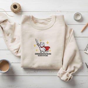 Könnte beinhalten: Cremefarbenes Sweatshirt mit einer Cartoon-Katze, die einen roten Umhang trägt und ein Schwert hält. Der Text "DISSERTATION SURVIVOR" steht darunter. Das Sweatshirt liegt auf einer weißen Holzoberfläche mit Kaffee, Stiften und Notizbuch.