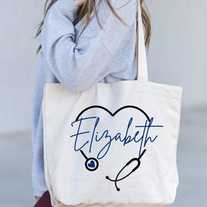 May include: Cream-colored tote bag featuring a black heart-shaped design with a stethoscope. The name "Elizabeth" is written in blue script within the heart. The bag is held by a person wearing a gray sweater.