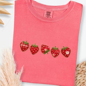 Puede incluir: Una camiseta de color coral con cinco diseños de fresas bordadas. Las fresas son rojas con hojas verdes y diferentes patrones, incluyendo lunares y cuadros. La camiseta tiene cuello redondo y una pequeña etiqueta.