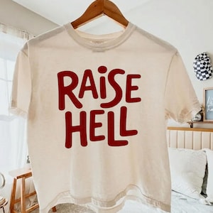 May include: Cream-colored t-shirt with the words "RAISE HELL" in bold, red letters. The shirt is hanging on a wooden hanger. The casual tee is a relaxed fit.