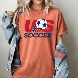Puede incluir: Una camiseta de manga corta color naranja quemado con un dise&ntilde;o gr&aacute;fico que presenta el texto rojo US, un bal&oacute;n de f&uacute;tbol blanco y negro que reemplaza la letra O, y el texto azul SOCCER debajo. El dise&ntilde;o incluye rayas azules con estrellas blancas.