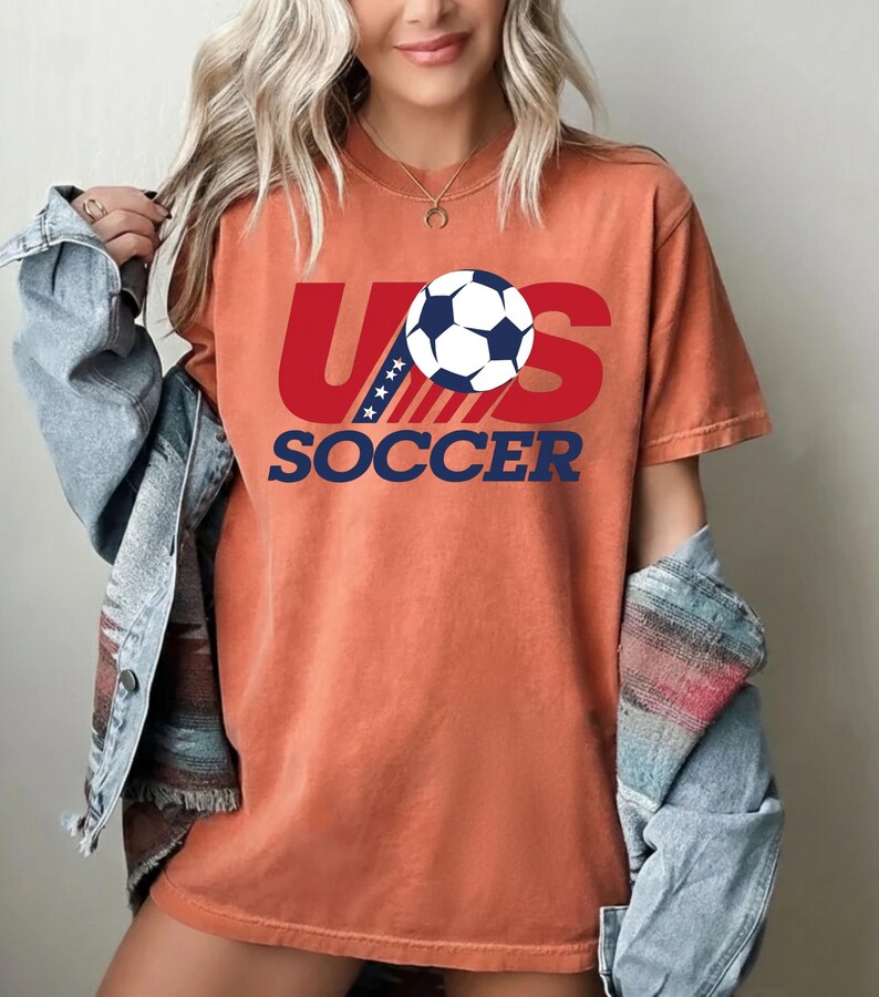 Puede incluir: Una camiseta de manga corta color naranja quemado con un dise&ntilde;o gr&aacute;fico que presenta el texto rojo US, un bal&oacute;n de f&uacute;tbol blanco y negro que reemplaza la letra O, y el texto azul SOCCER debajo. El dise&ntilde;o incluye rayas azules con estrellas blancas.
