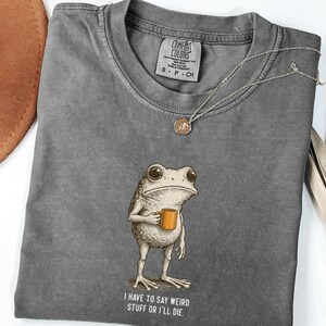 May include: A grey Comfort Colors t-shirt with a frog illustration holding a mug and the text "I have to say weird stuff or I'll die." A gold necklace and a brown hat are also visible. The t-shirt has a small label.