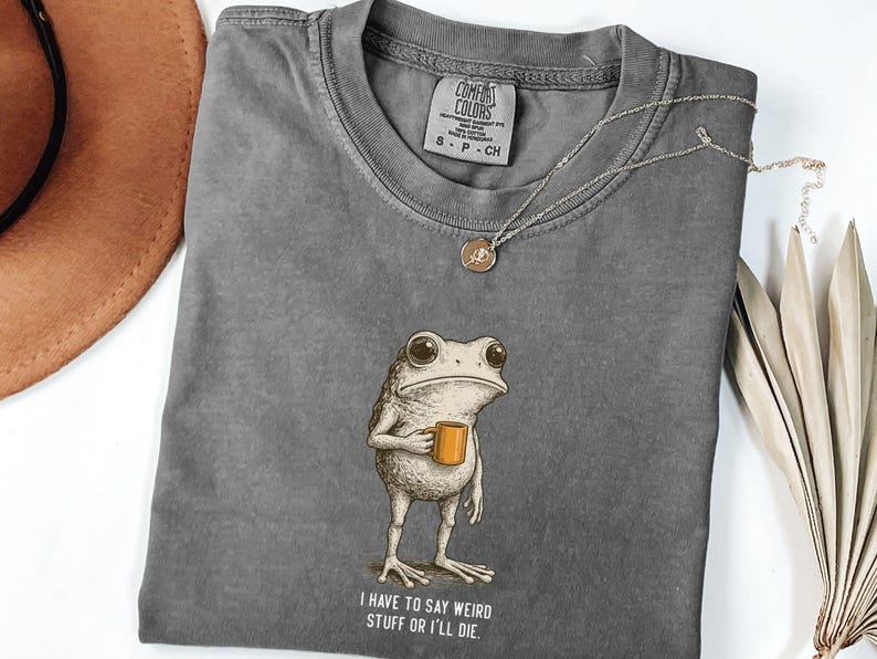 May include: A grey Comfort Colors t-shirt with a frog illustration holding a mug and the text "I have to say weird stuff or I'll die." A gold necklace and a brown hat are also visible. The t-shirt has a small label.