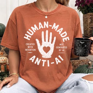 Może przedstawiać: T-shirt w kolorze rdzy z białą grafiką dłoni z wyciętym sercem pośrodku. Napis głosi: HUMAN-MADE, CREATED WITH INTENTION, NOT GENERATED BY AI oraz ANTI-AI. Wzór posiada postarzaną, przetartą teksturę nadruku.
