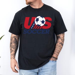 Puede incluir: Una camiseta negra de manga corta con un dise&ntilde;o gr&aacute;fico en el pecho. El dise&ntilde;o muestra el texto rojo US con estrellas blancas sobre una franja azul, un bal&oacute;n de f&uacute;tbol en blanco y negro, y la palabra SOCCER impresa en letras azules debajo.