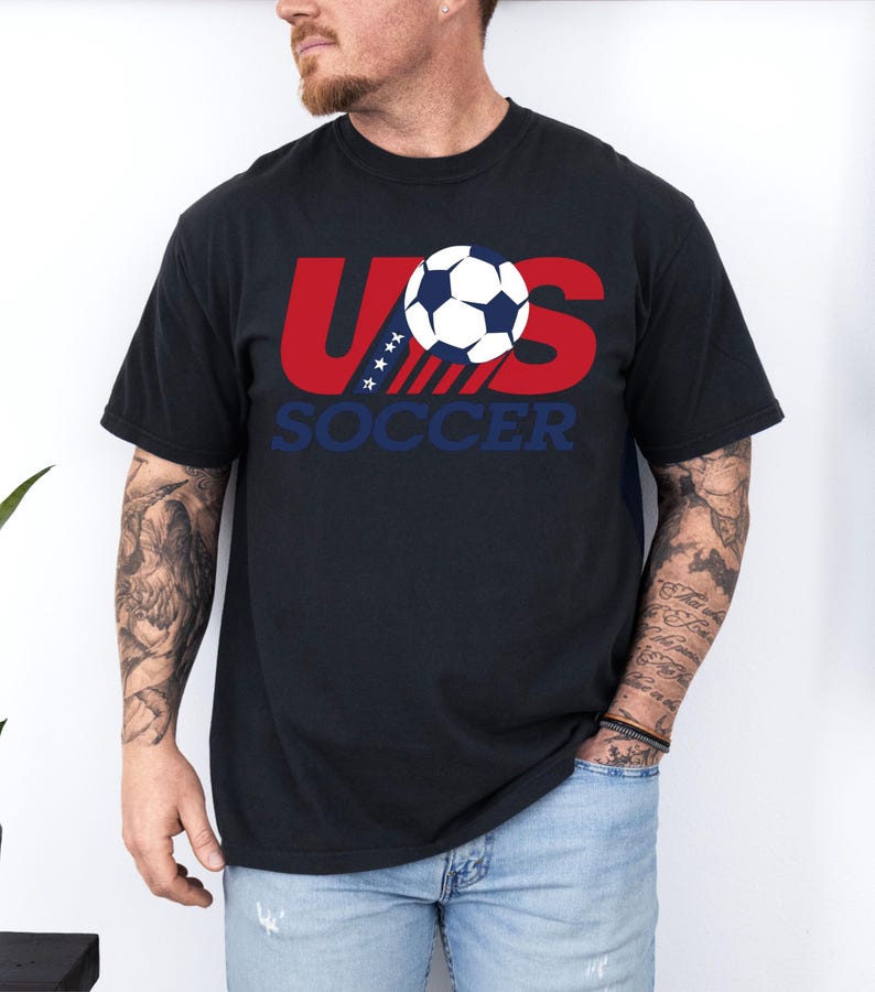 Puede incluir: Una camiseta negra de manga corta con un dise&ntilde;o gr&aacute;fico en el pecho. El dise&ntilde;o muestra el texto rojo US con estrellas blancas sobre una franja azul, un bal&oacute;n de f&uacute;tbol en blanco y negro, y la palabra SOCCER impresa en letras azules debajo.