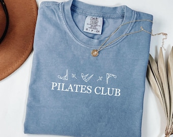 May include: A blue Comfort Colors t-shirt with the words "PILATES CLUB" in white. A gold necklace with a pendant rests on the shirt. A brown hat and dried plant are in the background.