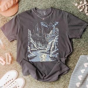 May include: Dark gray t-shirt featuring a fantasy landscape with a castle, river, and dragon. The design is in shades of blue, white, and gray. The shirt is surrounded by a peach-colored cap, white sneakers, and a light blue blanket with white hearts.