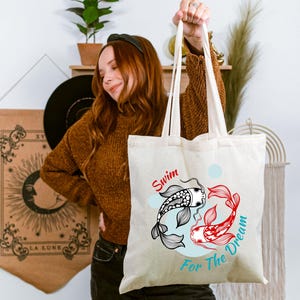 May include: Cream-colored tote bag featuring a koi fish design in black and red, with the words "Swim For The Dream" in red and blue. The bag has long handles and is held up by a person.