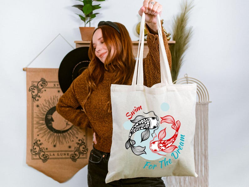 May include: Cream-colored tote bag featuring a koi fish design in black and red, with the words "Swim For The Dream" in red and blue. The bag has long handles and is held up by a person.