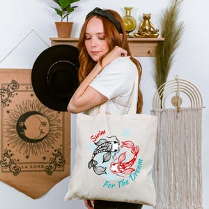 May include: Cream-colored tote bag featuring a koi fish design in black, red, and white, with the words "Swim For The Dream" in red and teal. The bag is held by a person.