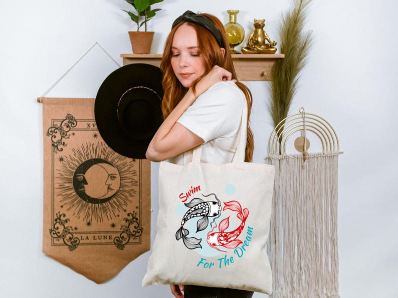 May include: Cream-colored tote bag featuring a koi fish design in black, red, and white, with the words "Swim For The Dream" in red and teal. The bag is held by a person.