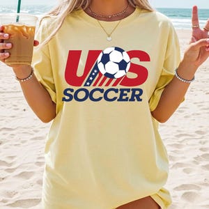 Puede incluir: Una camiseta de manga corta de color amarillo p&aacute;lido con un estampado gr&aacute;fico que dice US SOCCER en letras rojas y azul marino. El dise&ntilde;o incluye un bal&oacute;n de f&uacute;tbol blanco y negro que sustituye a la letra O y un patr&oacute;n de estrellas a rayas.