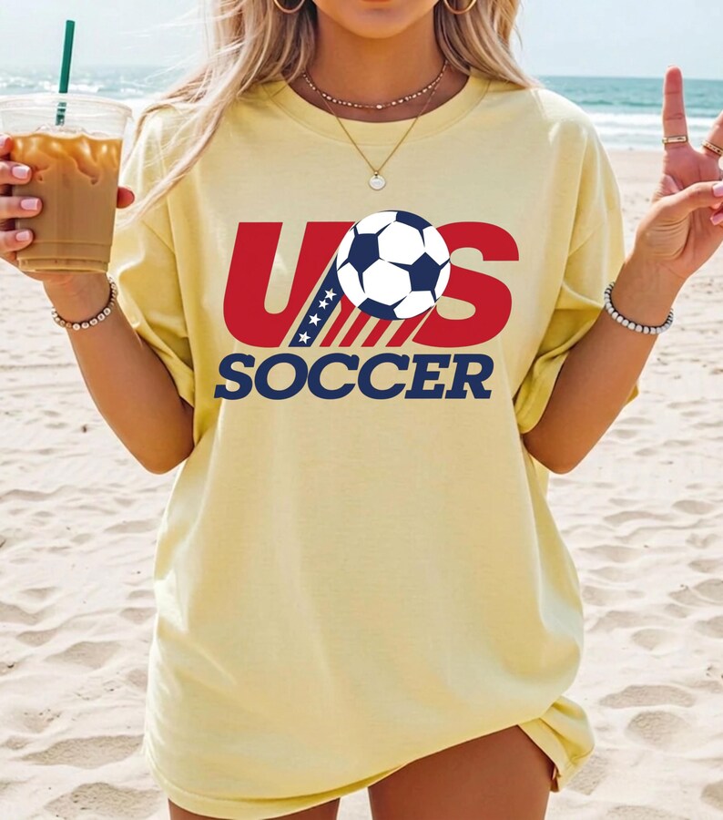 Puede incluir: Una camiseta de manga corta de color amarillo p&aacute;lido con un estampado gr&aacute;fico que dice US SOCCER en letras rojas y azul marino. El dise&ntilde;o incluye un bal&oacute;n de f&uacute;tbol blanco y negro que sustituye a la letra O y un patr&oacute;n de estrellas a rayas.