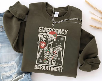 Puede incluir: Una sudadera verde oliva con las palabras "EMERGENCY DEPARTMENT" impresas en la parte delantera. El gráfico presenta un esqueleto con un gorro de Papá Noel y sosteniendo una taza con un símbolo médico. Una cadena de plata cuelga sobre la sudadera.