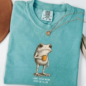 May include: A turquoise Comfort Colors t-shirt with a frog illustration holding a mug. The text "I HAVE TO SAY WEIRD STUFF OR I'LL DIE" is printed below the frog. A gold necklace and a brown hat are also visible.