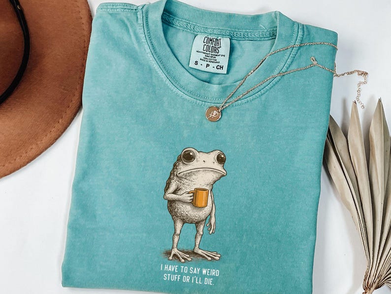 May include: A turquoise Comfort Colors t-shirt with a frog illustration holding a mug. The text "I HAVE TO SAY WEIRD STUFF OR I'LL DIE" is printed below the frog. A gold necklace and a brown hat are also visible.