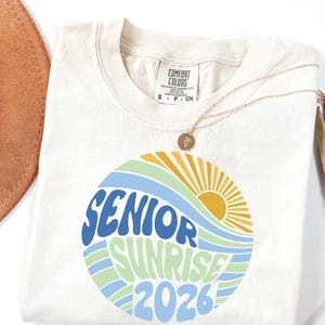 Puede incluir: Camiseta color crema con un dise&ntilde;o circular que dice "SENIOR SUNRISE 2026" en azul y verde, con un sol y olas amarillas. Un collar dorado con un colgante est&aacute; sobre la camiseta.