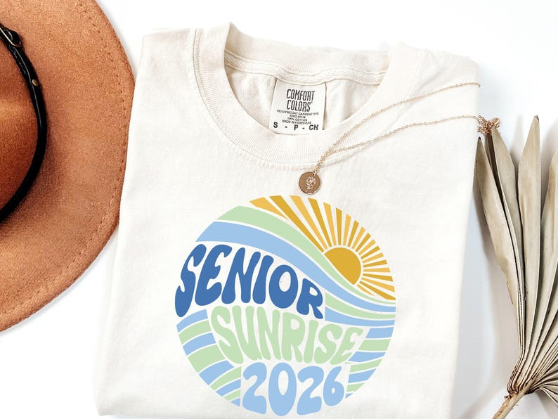 Puede incluir: Camiseta color crema con un dise&ntilde;o circular que dice "SENIOR SUNRISE 2026" en azul y verde, con un sol y olas amarillas. Un collar dorado con un colgante est&aacute; sobre la camiseta.