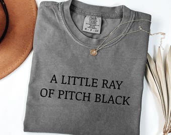 May include: A gray t-shirt with the text "A Little Ray of Pitch Black" in black. A gold pendant necklace and a brown hat are also visible. The shirt is folded and has a tag at the neckline.