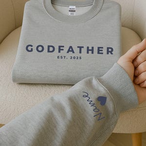 Pu&ograve; includere: Una felpa grigia con la scritta "GODFATHER" e l'anno "EST. 2025" stampati in blu navy. Il polsino della manica presenta un nome e un cuore blu. La felpa &egrave; piegata su una sedia color crema.