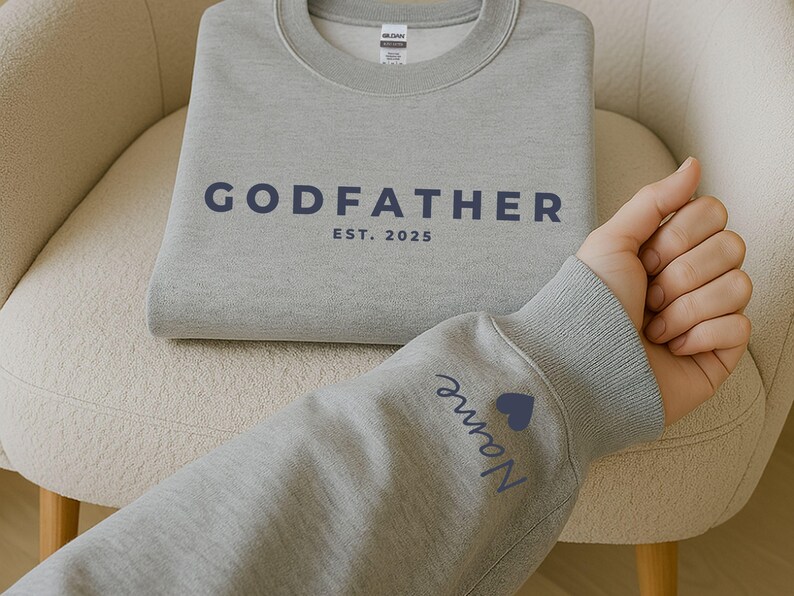 Pu&ograve; includere: Una felpa grigia con la scritta "GODFATHER" e l'anno "EST. 2025" stampati in blu navy. Il polsino della manica presenta un nome e un cuore blu. La felpa &egrave; piegata su una sedia color crema.