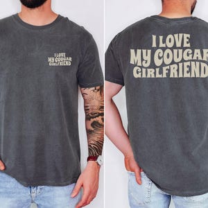 Może przedstawiać: Ciemnoszara koszulka z nadrukiem "I LOVE MY COUGAR GIRLFRIEND" z przodu i z tyłu w beżowej, retro czcionce. Koszulka ma okrągły dekolt i krótkie rękawy.