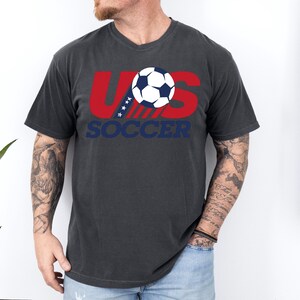Puede incluir: Una camiseta de manga corta gris carb&oacute;n con un gr&aacute;fico en el pecho. El dise&ntilde;o incluye el texto rojo US con cuatro estrellas blancas, un bal&oacute;n de f&uacute;tbol en blanco y negro, y la palabra SOCCER en letras may&uacute;sculas azules debajo.