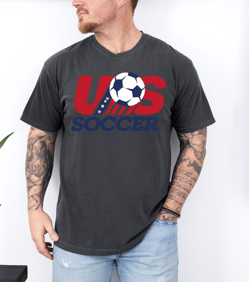 Puede incluir: Una camiseta de manga corta gris carb&oacute;n con un gr&aacute;fico en el pecho. El dise&ntilde;o incluye el texto rojo US con cuatro estrellas blancas, un bal&oacute;n de f&uacute;tbol en blanco y negro, y la palabra SOCCER en letras may&uacute;sculas azules debajo.