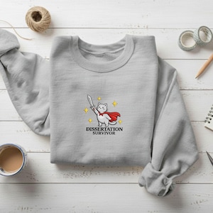 May include: A light gray sweatshirt featuring an embroidered cartoon cat wearing a red cape and holding a sword. The text "DISSERTATION SURVIVOR" is below the cat. The sweatshirt is on a white wooden surface with other stationery items.