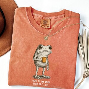 May include: Coral-coloured t-shirt with a frog graphic holding a mug, and the text "I have to say weird stuff or I'll die." A gold necklace and a brown hat are also visible.