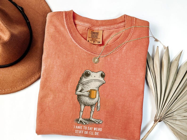 May include: Coral-coloured t-shirt with a frog graphic holding a mug, and the text "I have to say weird stuff or I'll die." A gold necklace and a brown hat are also visible.
