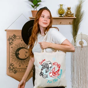 May include: Beige tote bag featuring a black and white koi fish and a red koi fish, with the words "Swim For The Dream" in red and teal. The bag is held by a person.