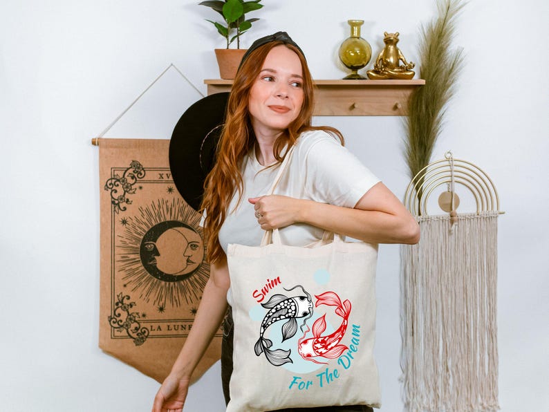 May include: Beige tote bag featuring a black and white koi fish and a red koi fish, with the words "Swim For The Dream" in red and teal. The bag is held by a person.