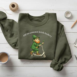 May include: Olive green sweatshirt with the phrase "Off to commit Irish foolery" above a cartoon leprechaun riding a scooter. The leprechaun is wearing green and holding a shamrock. The sweatshirt is on a white wooden surface.