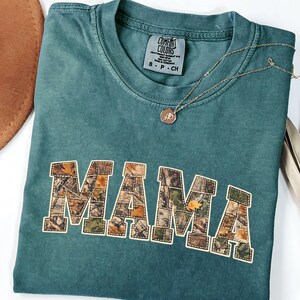 Puede incluir: Una camiseta Comfort Colors verde azulado con la palabra "MAMA" en un patrón de camuflaje. Un collar dorado con un colgante está sobre la camiseta. Un sombrero marrón y hojas de palma secas están en el fondo.