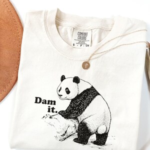 Peut inclure: T-shirt crème avec un motif d'ours panda noir et blanc et le texte "Dam it." Un collier en or avec un pendentif rond est posé sur le t-shirt. Un chapeau marron et une plante séchée sont en arrière-plan.