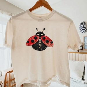 May include: Cream-colored t-shirt featuring a large ladybug applique. The ladybug has a black body with red and black polka dot wings. The wings have a plaid pattern. The antennae are black, and the design is stitched with a cross-stitch pattern.
