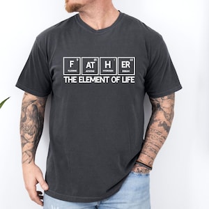 Puede incluir: Una camiseta gris oscuro con un gráfico blanco de la palabra "FATHER" deletreada usando símbolos de elementos químicos. El texto "THE ELEMENT OF LIFE" está impreso debajo del gráfico.