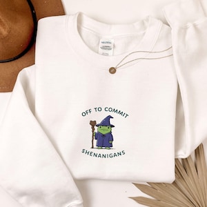 May include: White sweatshirt featuring a cartoon frog wizard. The frog wears a blue wizard hat and robe, holding a staff. Text reads "Off to Commit Shenanigans". A gold necklace and brown hat are also visible.