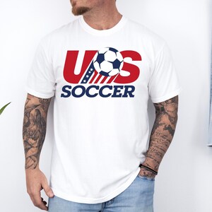 Puede incluir: Una camiseta blanca de manga corta con un dise&ntilde;o gr&aacute;fico en el pecho. El gr&aacute;fico muestra el texto US en letras rojas grandes con un patr&oacute;n de rayas azules y blancas, un bal&oacute;n de f&uacute;tbol en blanco y negro y la palabra SOCCER en azul.