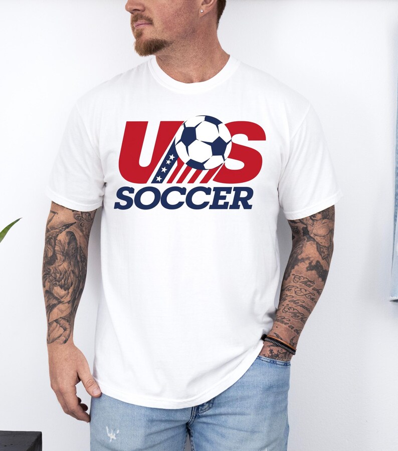 Puede incluir: Una camiseta blanca de manga corta con un dise&ntilde;o gr&aacute;fico en el pecho. El gr&aacute;fico muestra el texto US en letras rojas grandes con un patr&oacute;n de rayas azules y blancas, un bal&oacute;n de f&uacute;tbol en blanco y negro y la palabra SOCCER en azul.