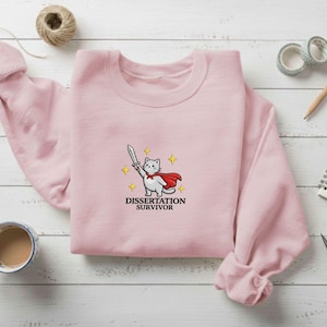 May include: A light pink sweatshirt featuring an embroidered design of a white cat wearing a red cape and holding a sword, with the text "Dissertation Survivor" below. The sweatshirt is displayed on a wooden surface with other stationery items.