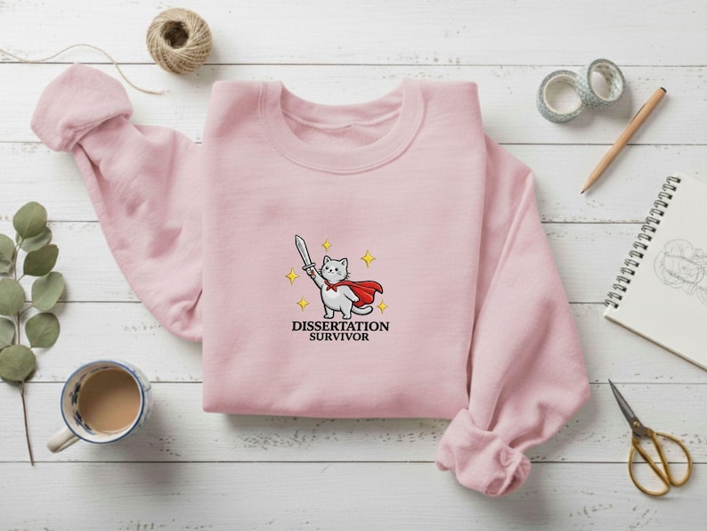 May include: A light pink sweatshirt featuring an embroidered design of a white cat wearing a red cape and holding a sword, with the text "Dissertation Survivor" below. The sweatshirt is displayed on a wooden surface with other stationery items.