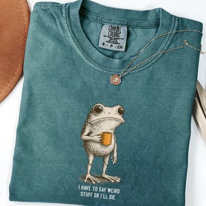 May include: Teal Comfort Colors t-shirt with a frog graphic holding a mug, and the text "I HAVE TO SAY WEIRD STUFF OR I'LL DIE." A gold necklace and a brown hat are also visible.
