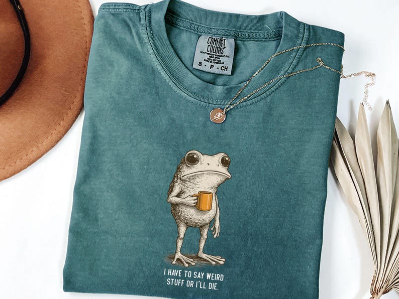 May include: Teal Comfort Colors t-shirt with a frog graphic holding a mug, and the text "I HAVE TO SAY WEIRD STUFF OR I'LL DIE." A gold necklace and a brown hat are also visible.