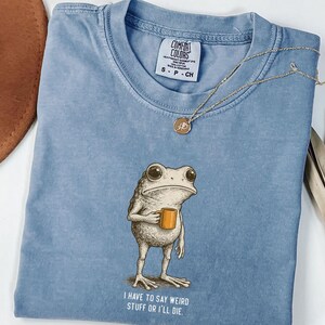 May include: A pale blue Comfort Colors t-shirt with a frog illustration holding a mug. The text reads "I have to say weird stuff or I'll die". A gold pendant necklace is also visible. The shirt is size S-P-CH.